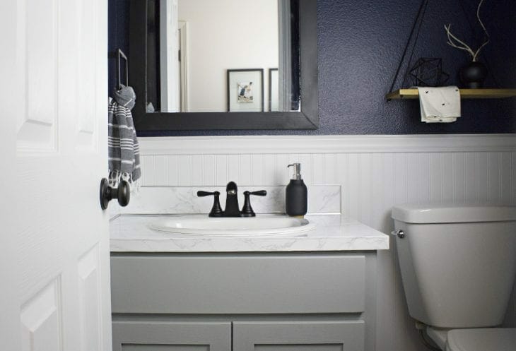 This moody and dark bathroom makeover reveal is STUNNING! Between the navy wall color, updated mirror, faux countertops, and matte black accessories, this whole small powder room turned out amazing.