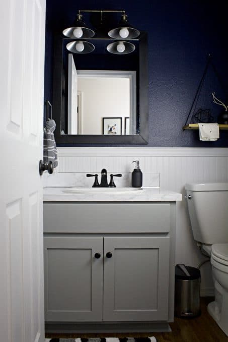 This moody and dark bathroom makeover reveal is STUNNING! Between the navy wall color, updated mirror, faux countertops, and matte black accessories, this whole small powder room turned out amazing.