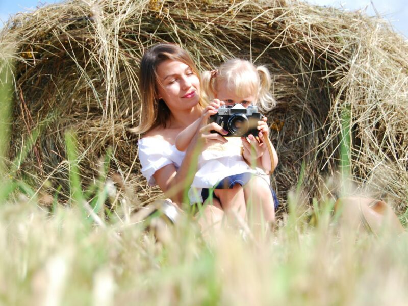 woman in white tank top holding black dslr camera sitting on brown grass field during daytime