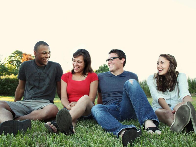 A group of young adults smiling while sitting on grass in a sunny park, enjoying friendship and leisure.