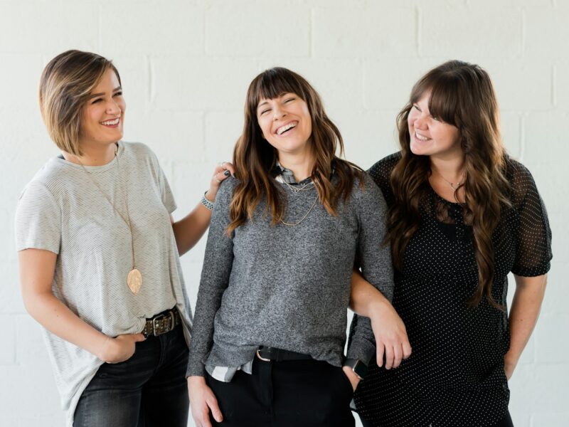 3 women smiling and standing beside white wall