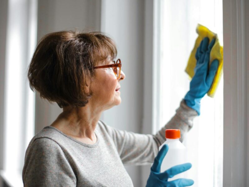 Elderly woman cleansing window indoors using gloves and cleaner.