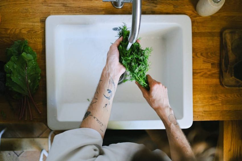 Tattooed hands washing fresh herbs in a kitchen sink, highlighting daily culinary routines.