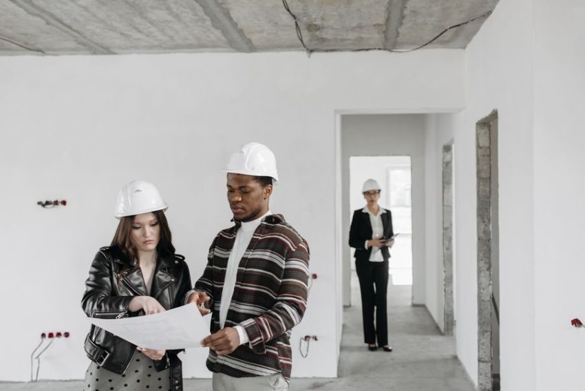 A couple wearing hard hats examines building plans inside an unfinished property with a realtor observing.