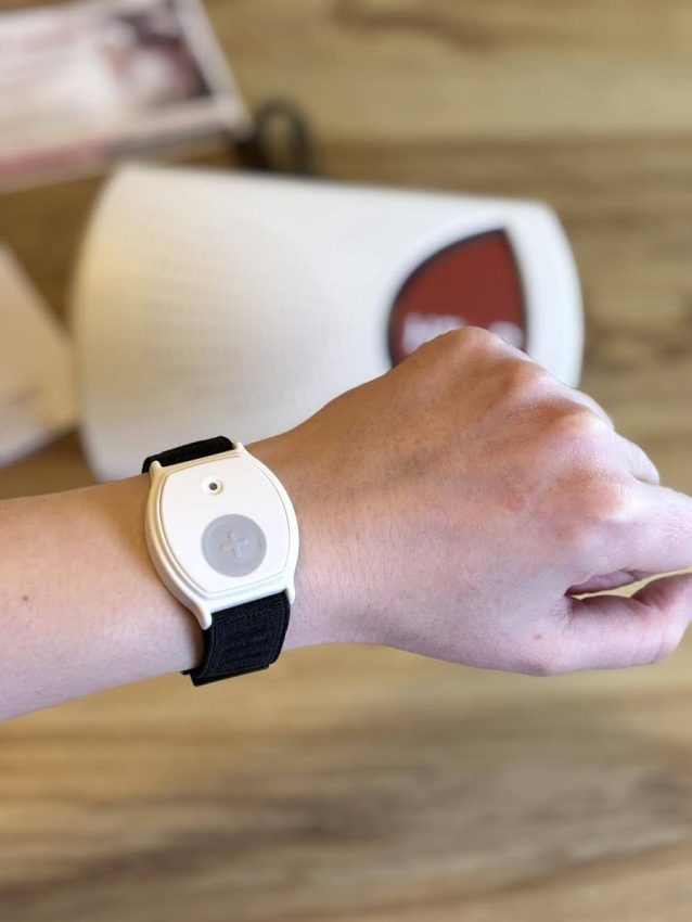 White medical alert button on a black adjustable fabric strap worn on a person's wrist