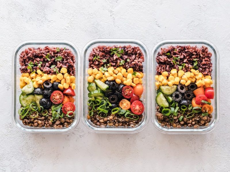 Three glass containers filled with rice, corn, olives, tomatoes, cucumbers, and lentils