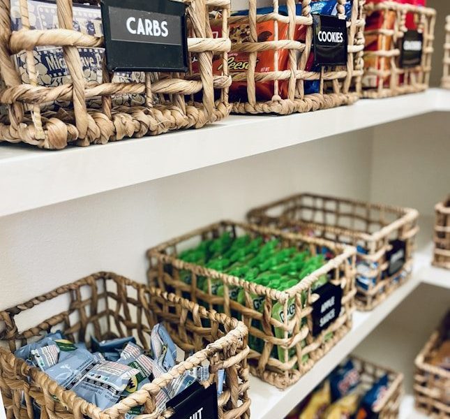Woven baskets with labels on white pantry shelves filled with various snacks